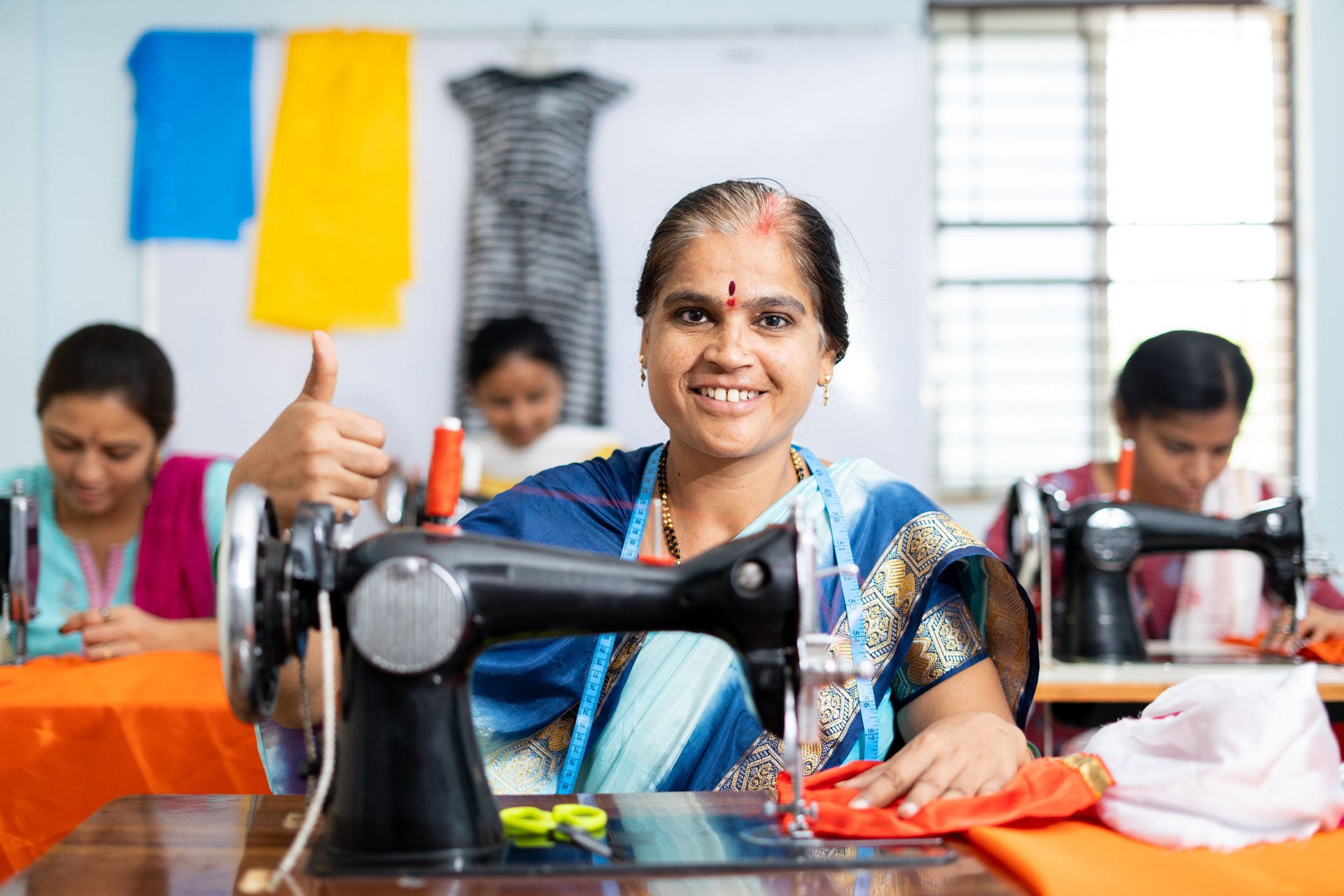 Happy smiling woman showing thumbs up sign by looking camera while working at garments sewing maching - concept of employment, job and successful business