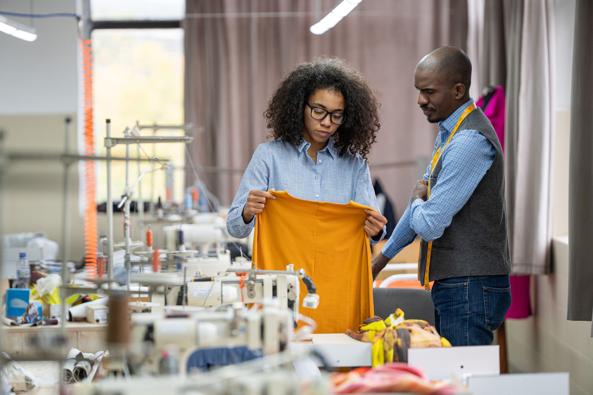 Focused young african-american female tailor in a design studio showing a textile product to a male designer, having discussion among colleagues, fashion ideas exchange and teamwork